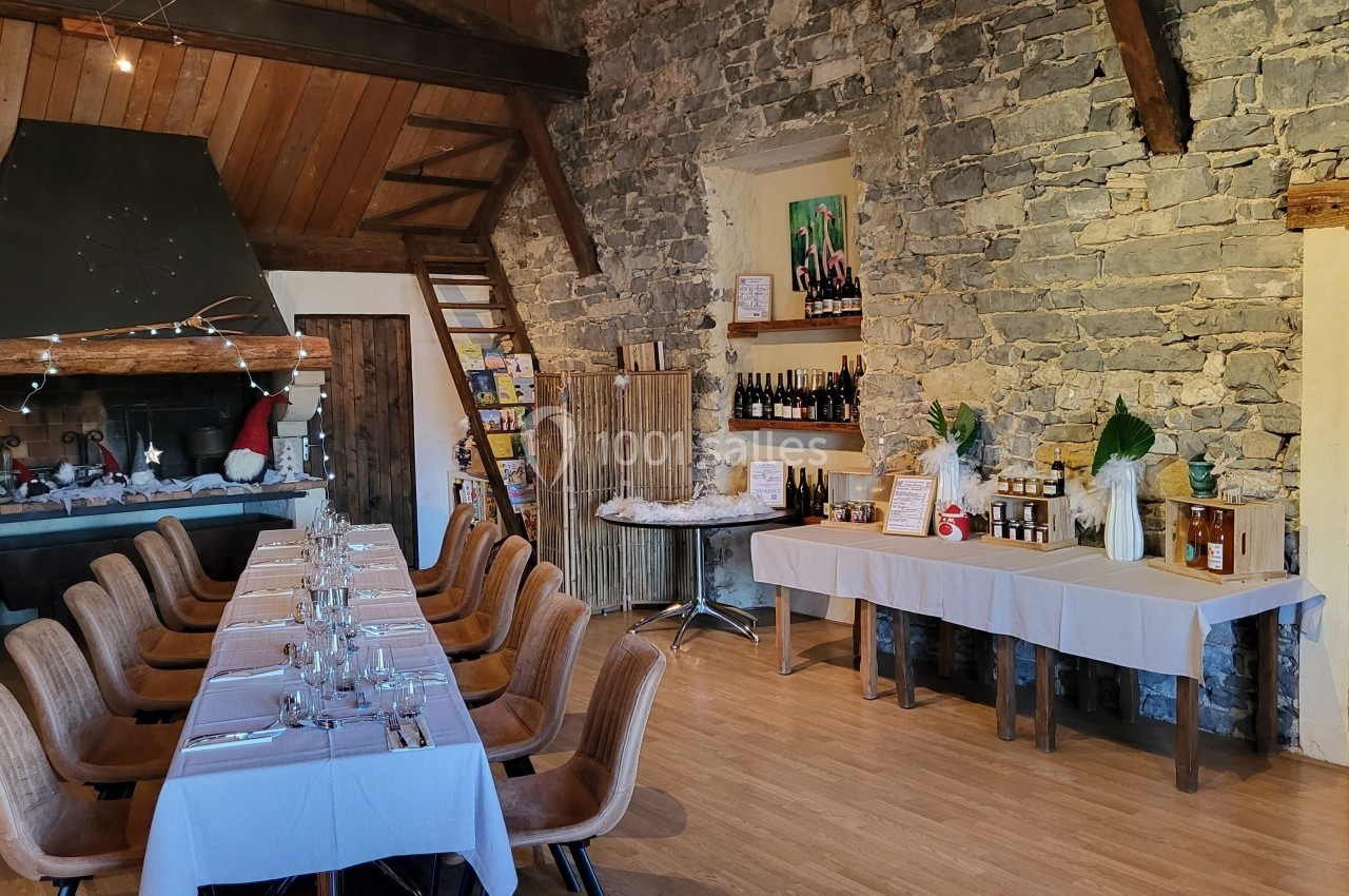 Salle rustique avec murs en pierre, grande table dressée, cheminée décorée et tables présentant des produits locaux.
