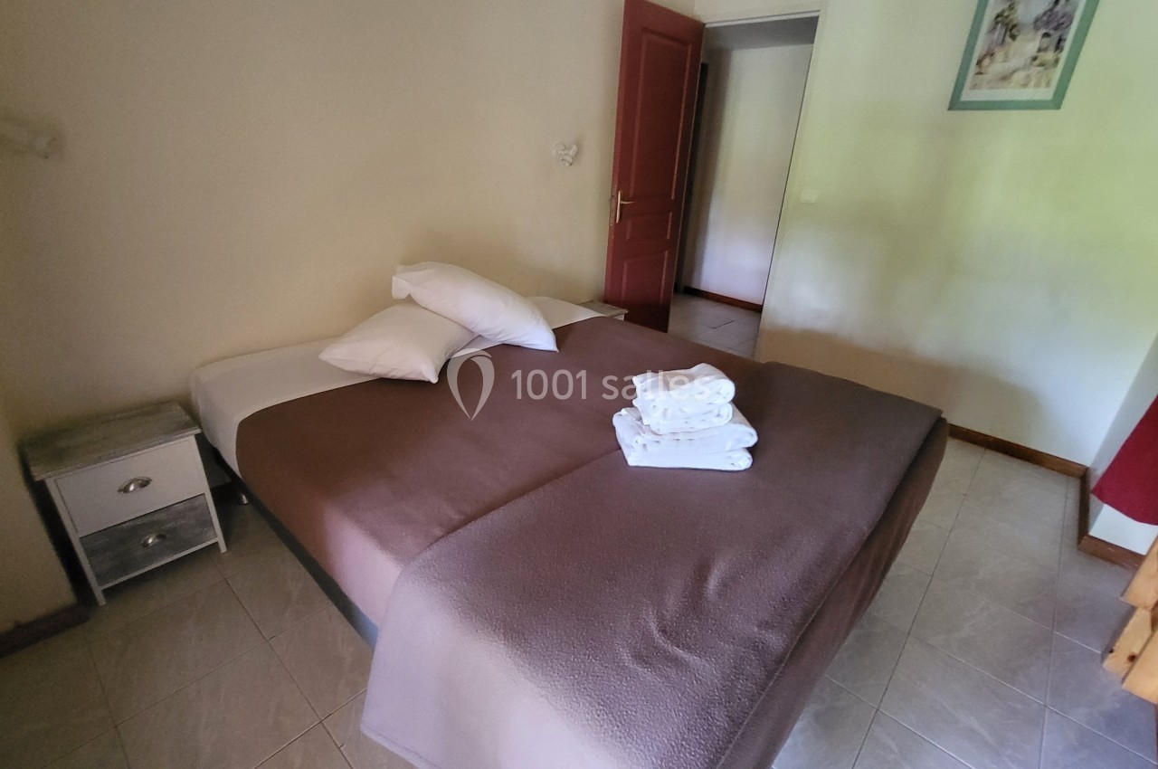 Lit double avec draps bruns, oreillers blancs et serviettes pliées, dans une chambre lumineuse avec porte rouge ouverte.