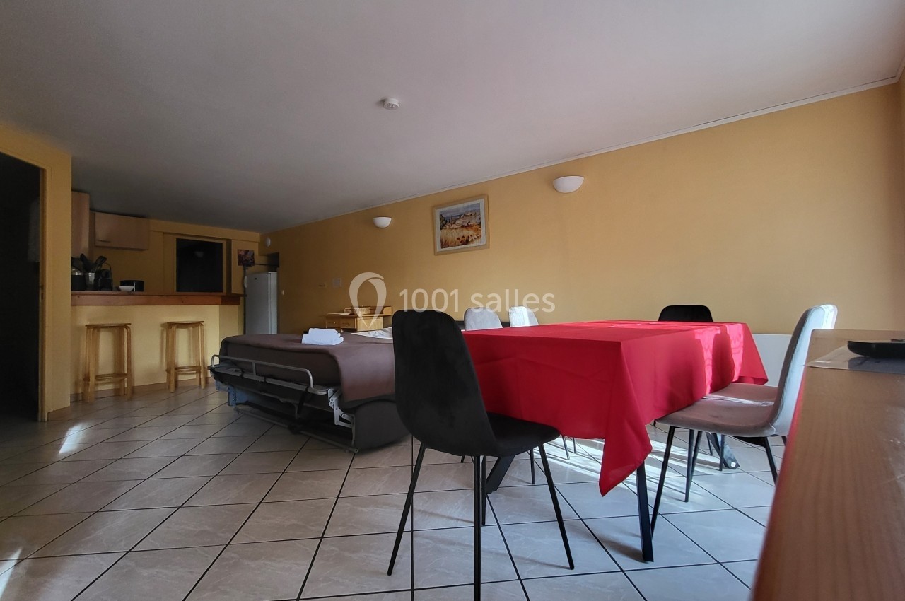Salle à manger avec table recouverte d'une nappe rouge, chaises noires et blanches, cuisine ouverte et canapé-lit en arrière…