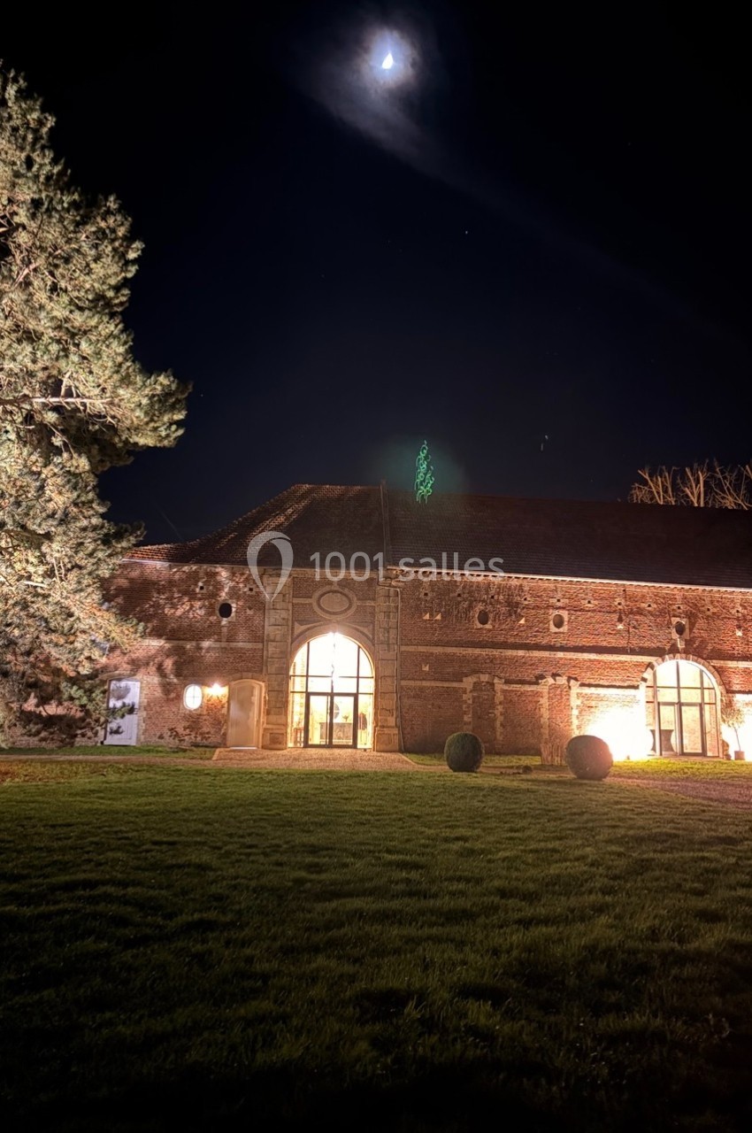 Bâtiment en briques éclairé la nuit, avec une pelouse au premier plan et un arbre sous un ciel étoilé.