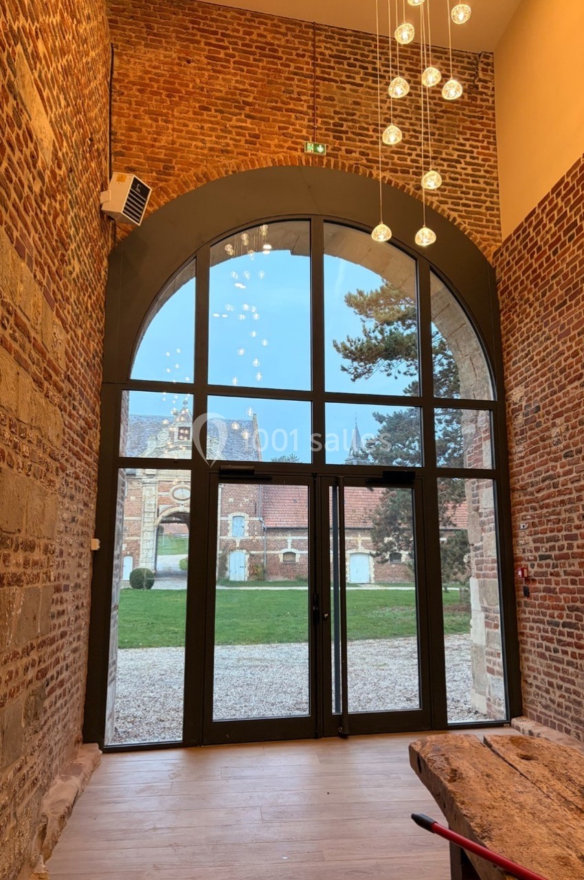Grande baie vitrée en arche dans un mur en briques, donnant sur une cour avec des bâtiments anciens et un arbre.