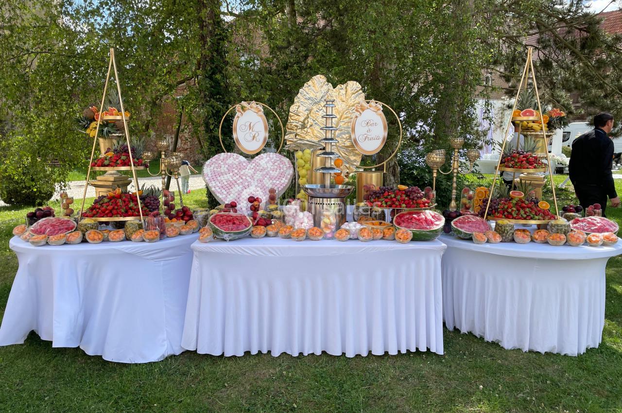Buffet extérieur avec tables garnies de fruits variés, fontaine à chocolat et décorations dorées sous des arbres.