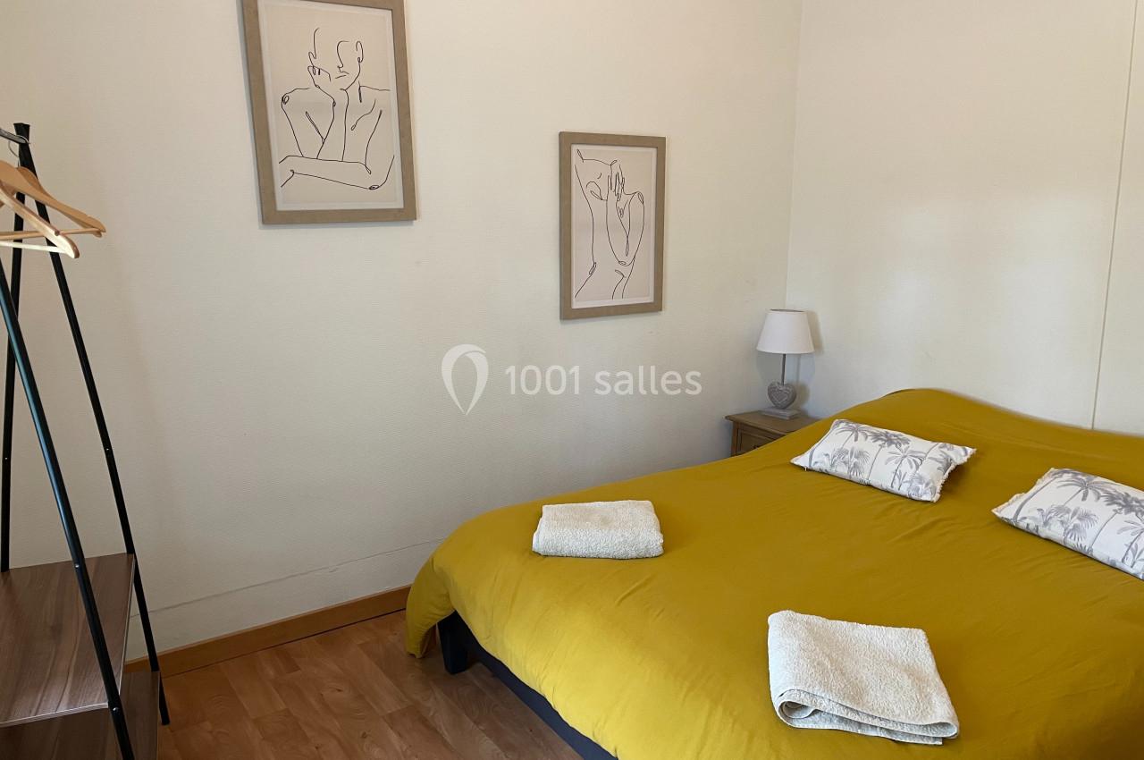 Chambre avec un lit double couvert d'une housse jaune, deux serviettes pliées et des dessins encadrés au mur.