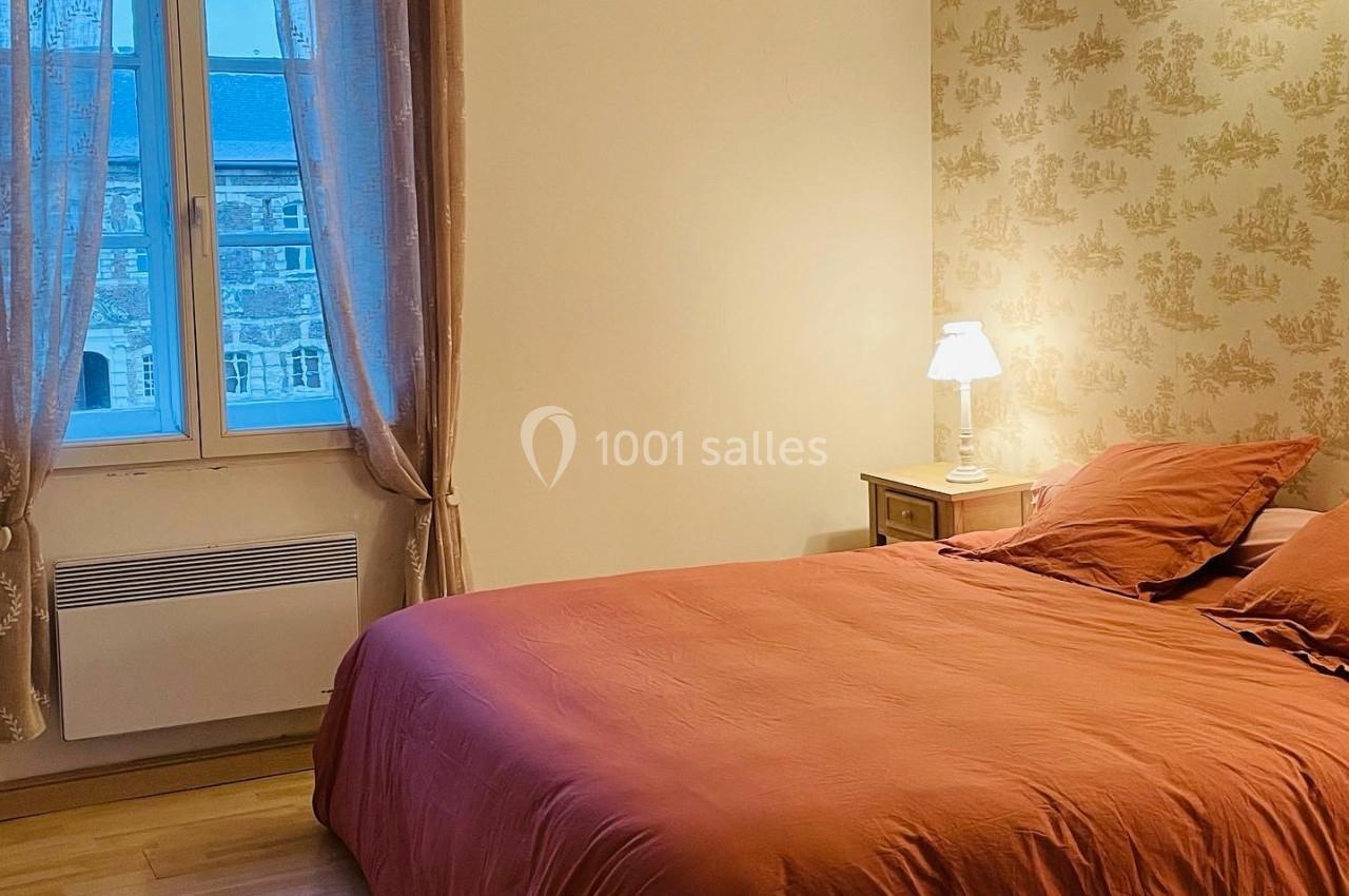 Chambre lumineuse avec un lit double aux draps orange, une lampe de chevet et une fenêtre donnant sur des bâtiments.