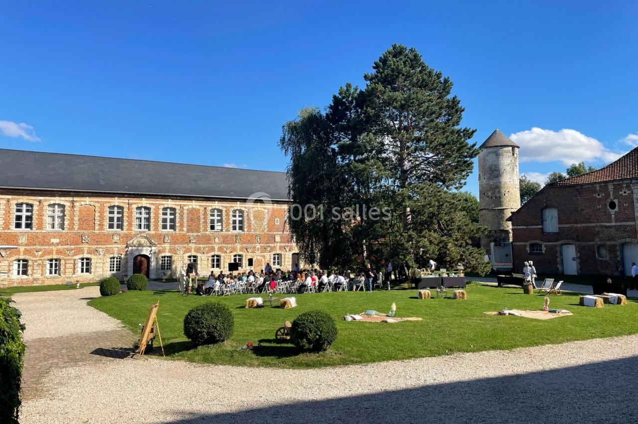 Cour d'une ancienne bâtisse en briques avec pelouse, arbres, tour ronde et invités rassemblés pour un événement.