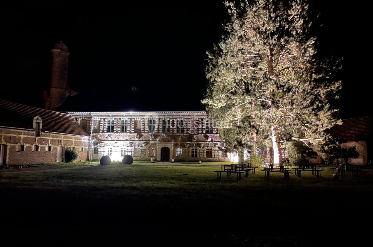 Façade d'un bâtiment ancien éclairé la nuit, entouré d'arbres et de tables sur une pelouse.