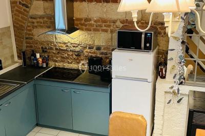 Miniature Cuisine du gîte Pièce rustique avec murs en pierre, plafond en bois, réservoir métallique rouge et mobilier simple.