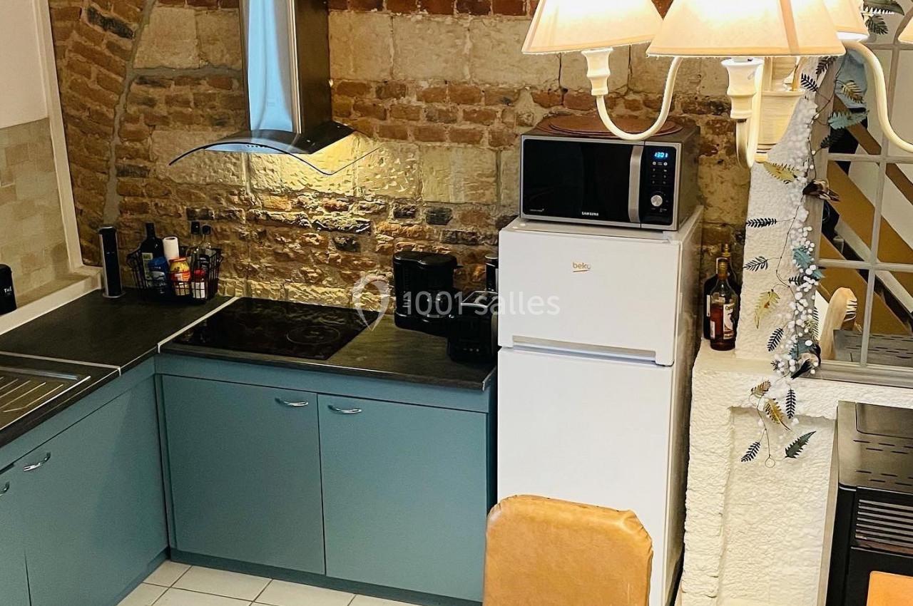 Cuisine avec murs en briques apparentes, meubles bleu-gris, table ronde en bois, réfrigérateur blanc et éclairage suspendu.