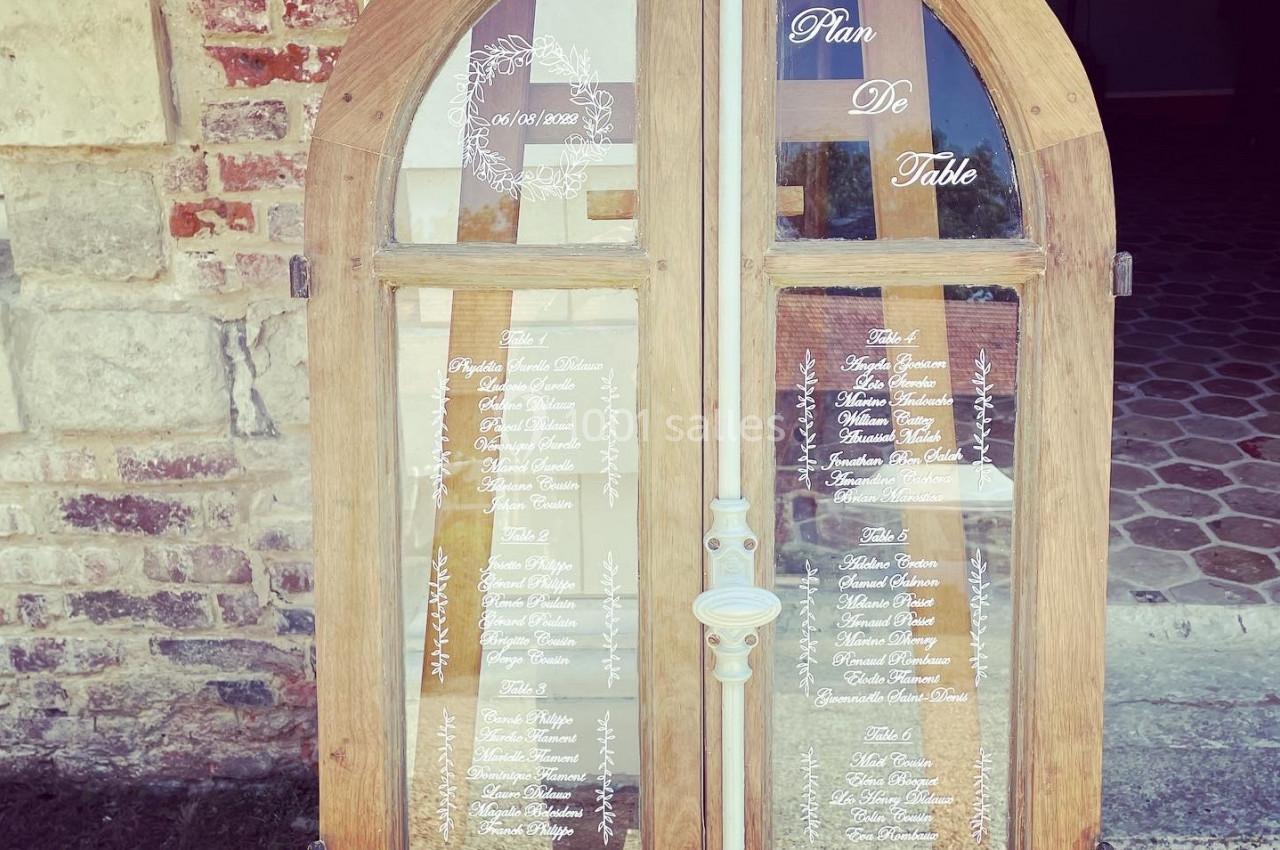 Plan de table inscrit sur des vitres d'une porte en bois, posé devant un mur en briques et une entrée pavée.