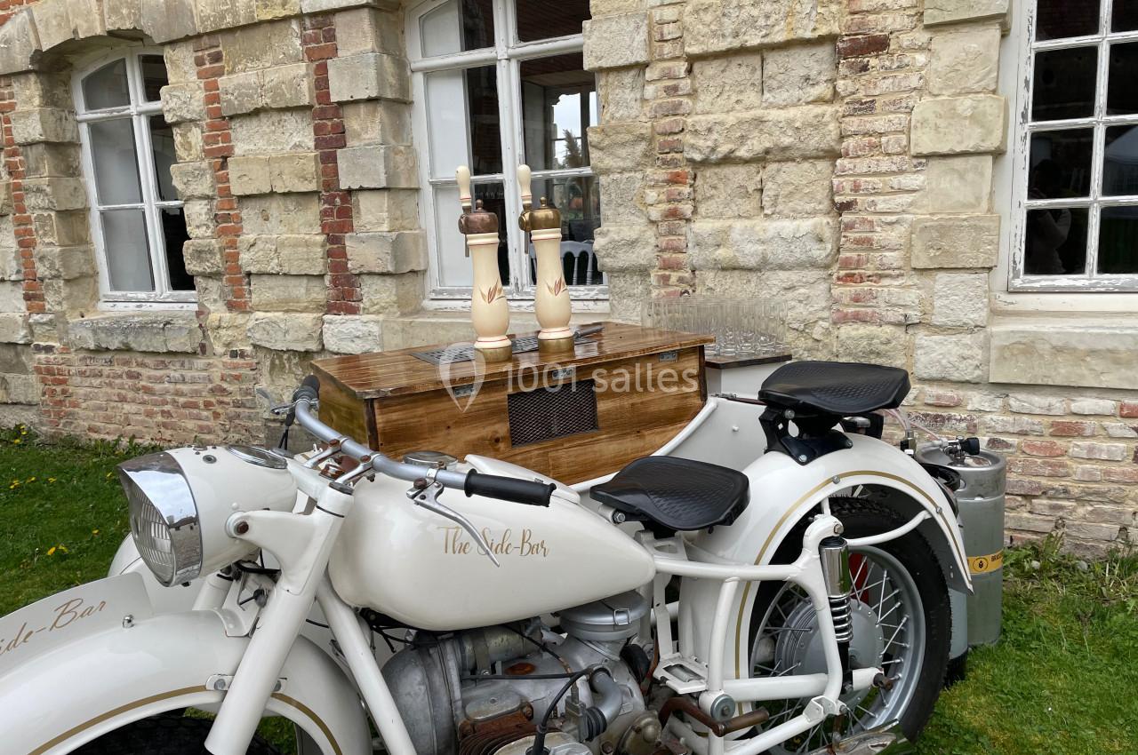 Moto vintage blanche avec side-car aménagé en bar mobile, stationnée devant un bâtiment en briques et pierres.