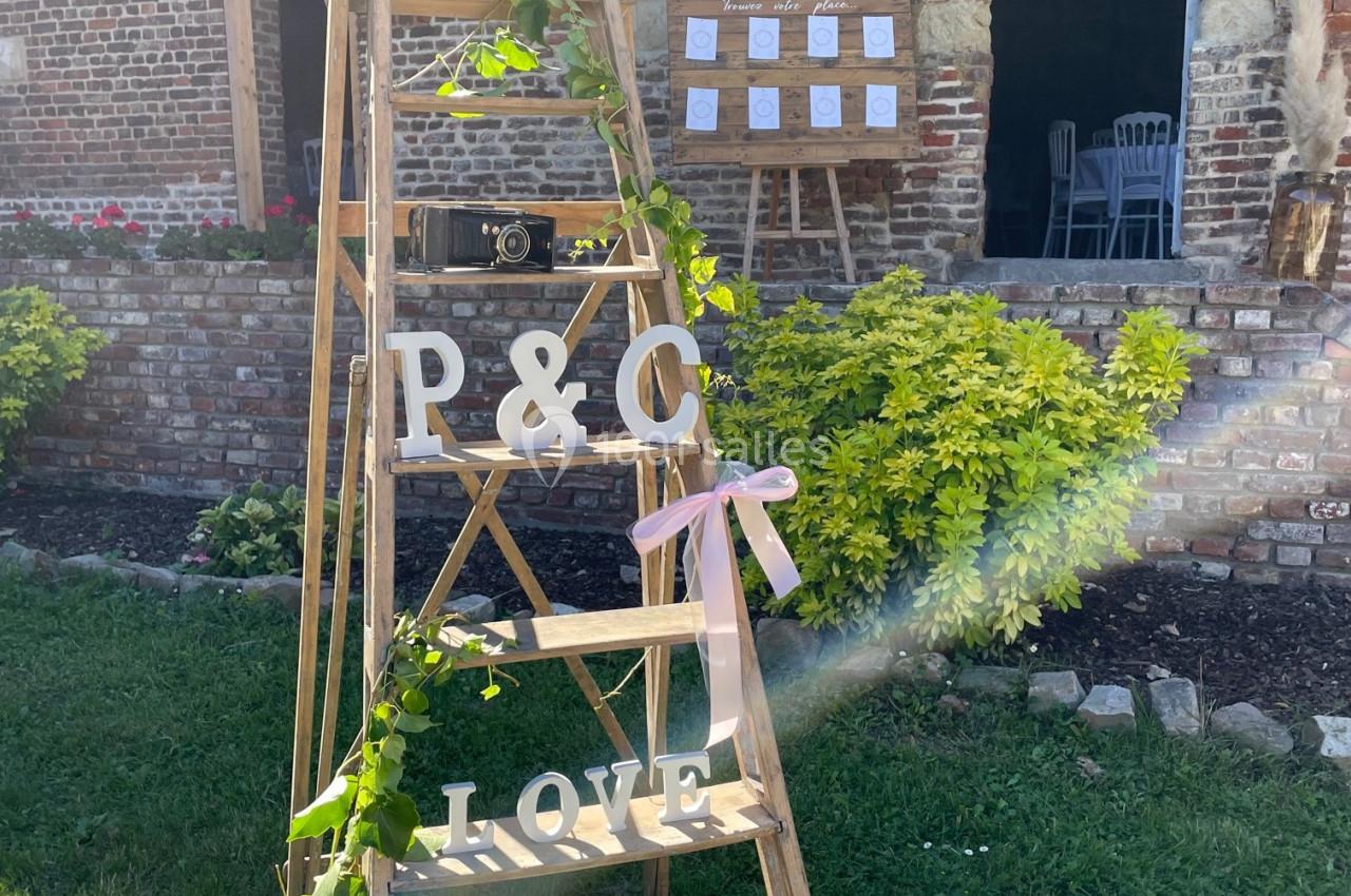 Échelle en bois décorée de feuillage, lettres ’P&C’ et ’LOVE’, devant un mur en briques avec un panneau en arrière-plan.