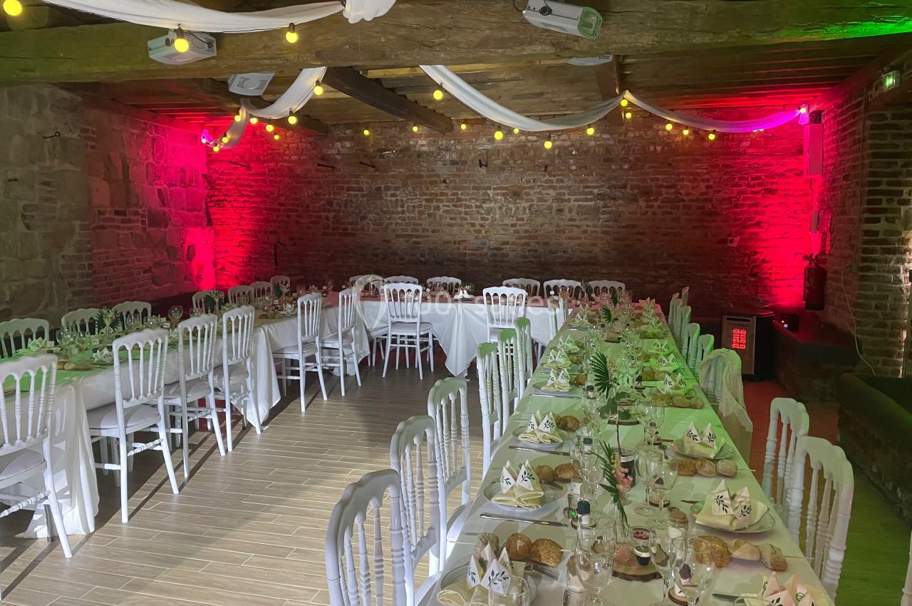 Salle en pierre avec tables dressées pour un événement, décorée de guirlandes lumineuses et de nappes blanches.