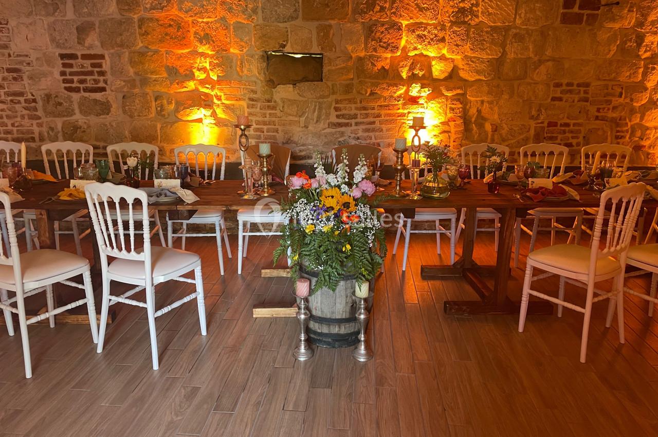 Salle en pierre éclairée d'une lumière chaude, avec des tables décorées et des chaises blanches disposées autour.