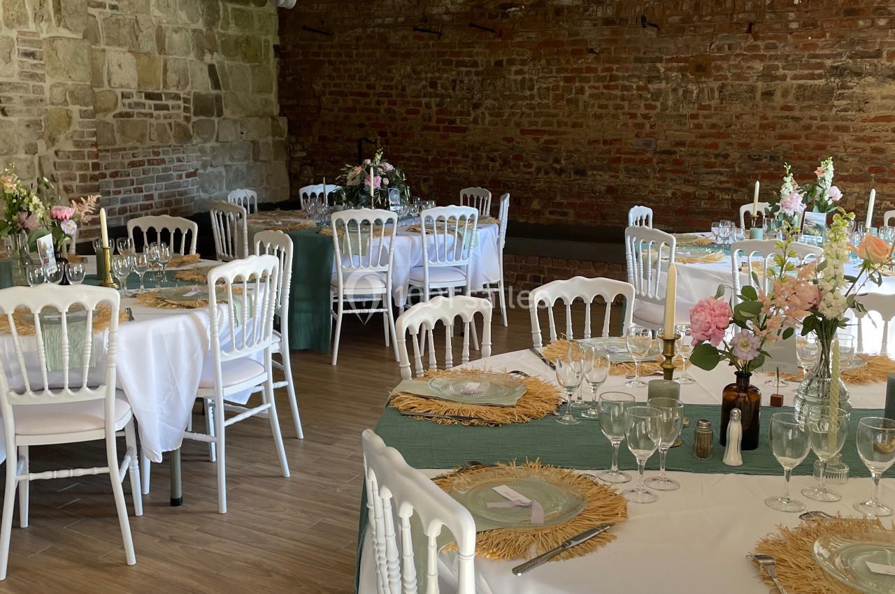 Salle de réception avec tables rondes dressées, nappes blanches, chaises blanches et décorations florales.