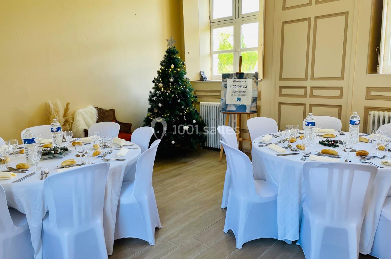 Salle décorée pour un repas, avec tables rondes dressées, nappes blanches et un sapin de Noël illuminé.