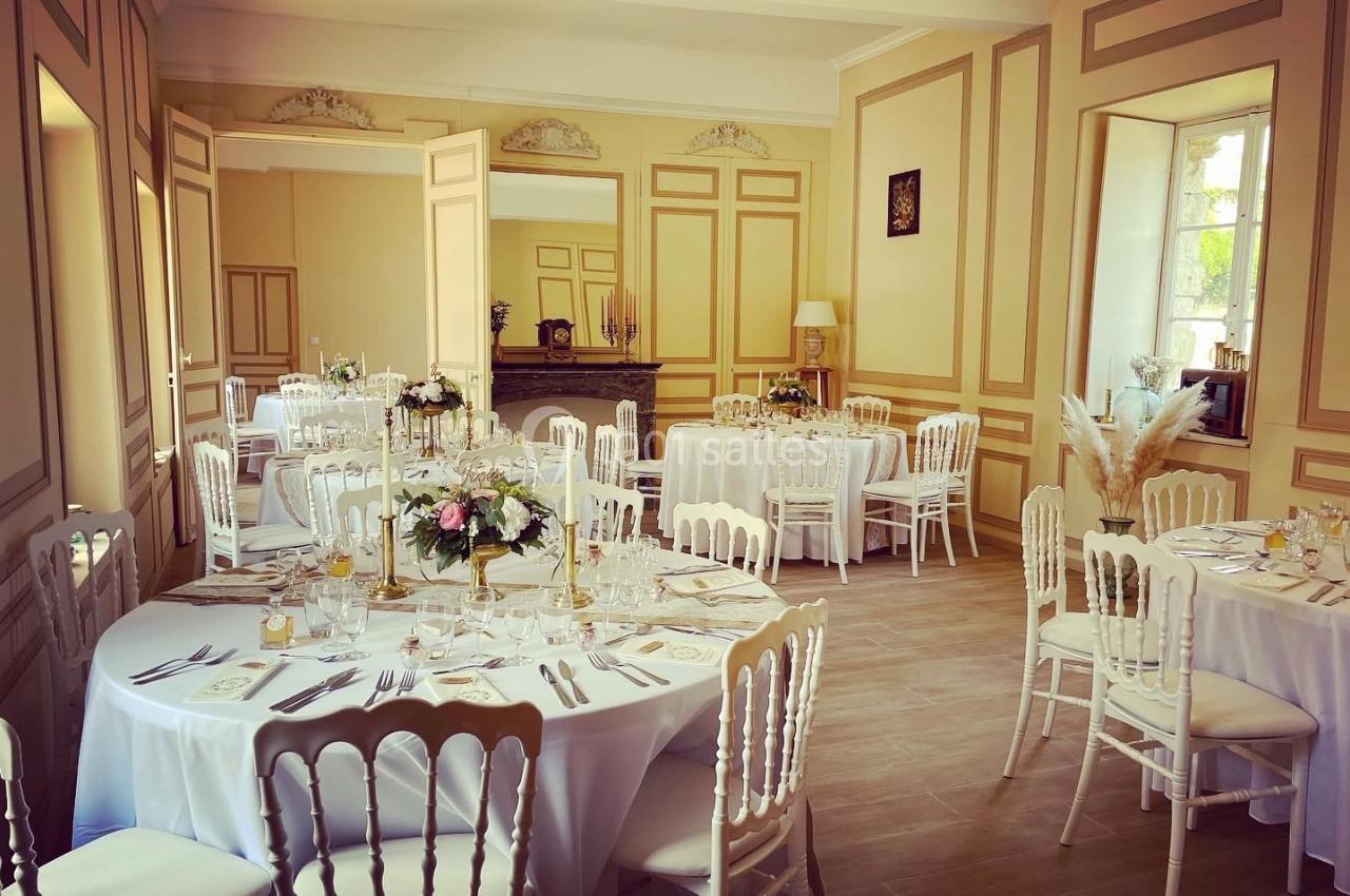 Salle élégante avec tables rondes dressées pour un événement, décorée de fleurs et baignée de lumière naturelle.