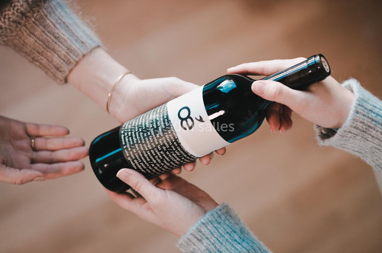 Deux mains tendent une bouteille de vin à une troisième main dans un environnement intérieur lumineux.