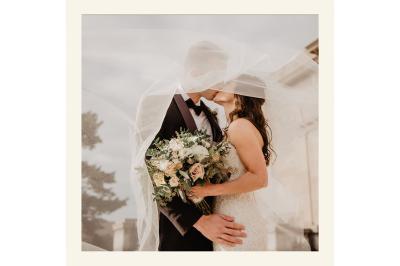 Un couple en tenue de mariage s'embrasse sous un voile, tenant un bouquet de fleurs.