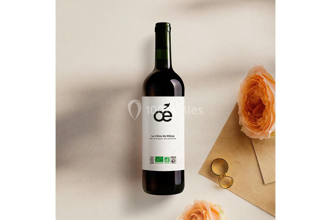 Bouteille de vin rouge posée sur une surface beige, accompagnée de roses orangées et d'une paire de lunettes dorées.