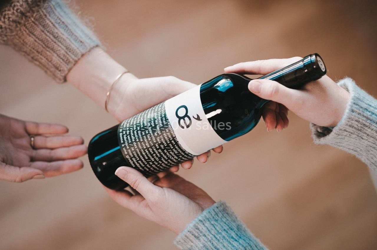 Deux mains tendent une bouteille de vin à une troisième main, sur un fond flou en intérieur.