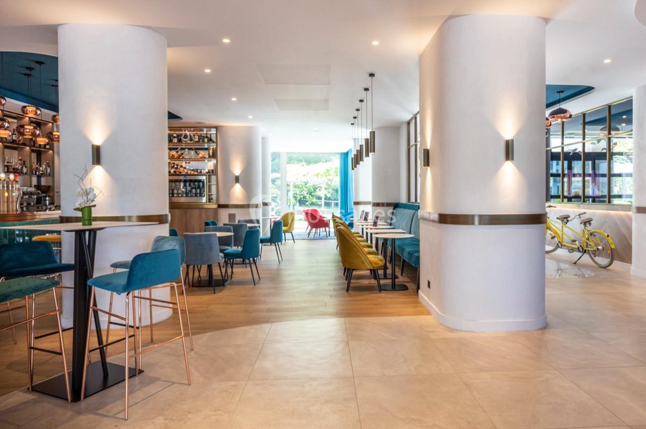Intérieur lumineux d'un café moderne avec tables, chaises colorées, bar et grandes baies vitrées donnant sur l'extérieur.