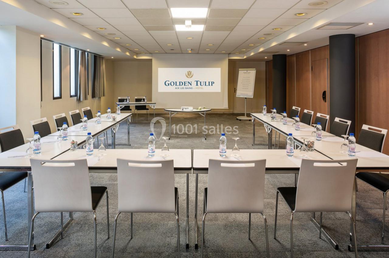 Salle de réunion lumineuse avec tables disposées en U, chaises blanches et écran de projection.