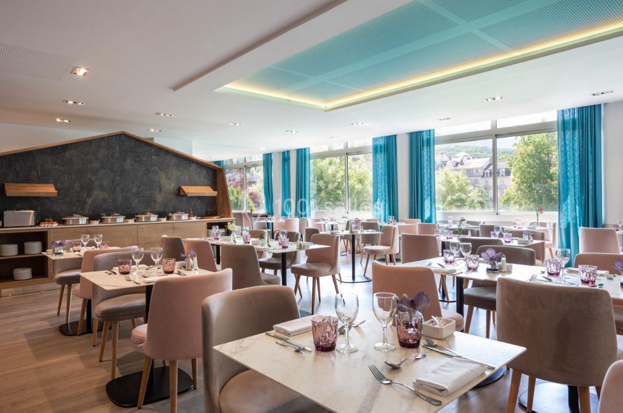 Salle de restaurant lumineuse avec tables dressées, chaises rembourrées et grandes fenêtres donnant sur un espace arboré.