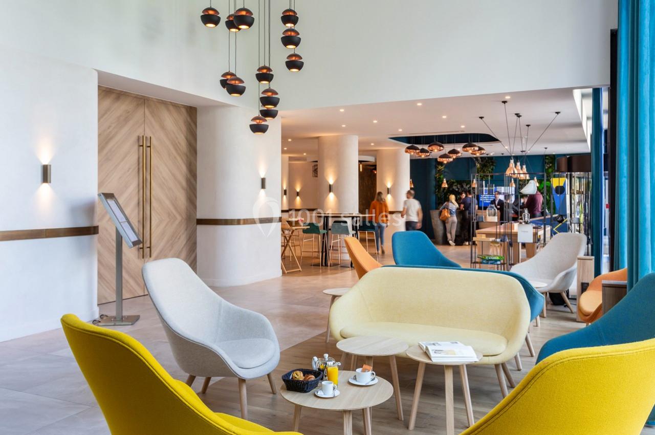 Espace lounge moderne avec fauteuils colorés, tables basses et éclairage suspendu, donnant sur une zone animée en arrière…