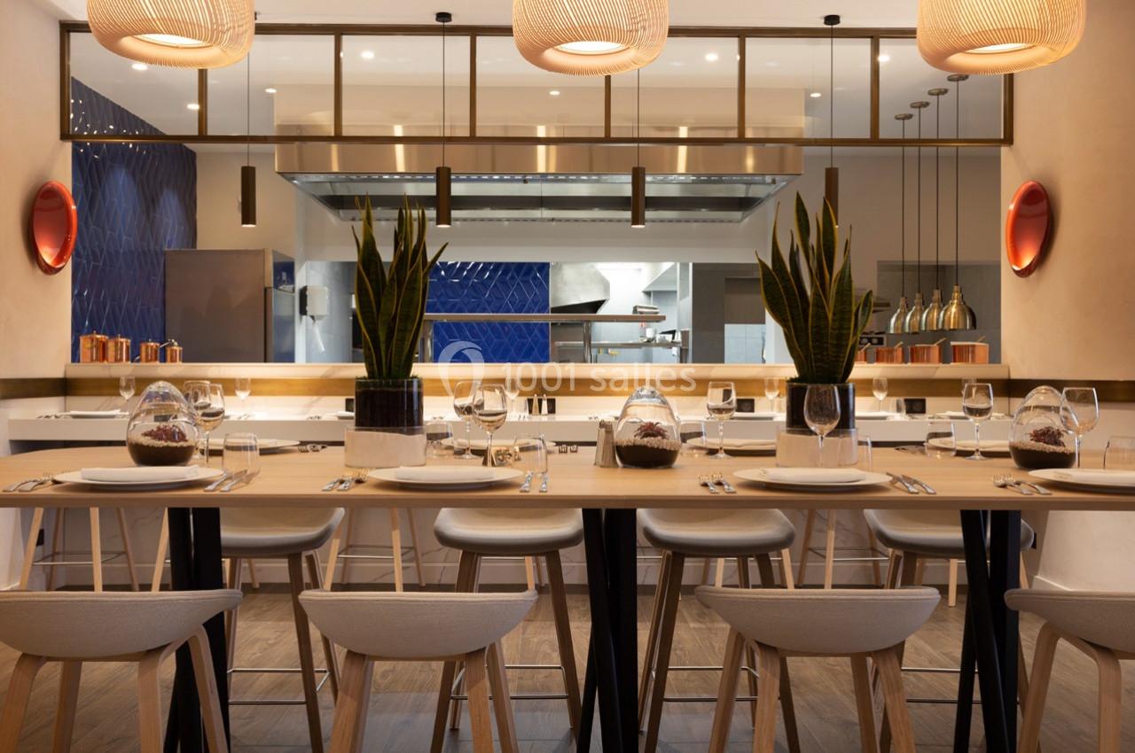 Salle de restaurant moderne avec tables dressées, plantes décoratives et vue sur une cuisine ouverte en arrière-plan.