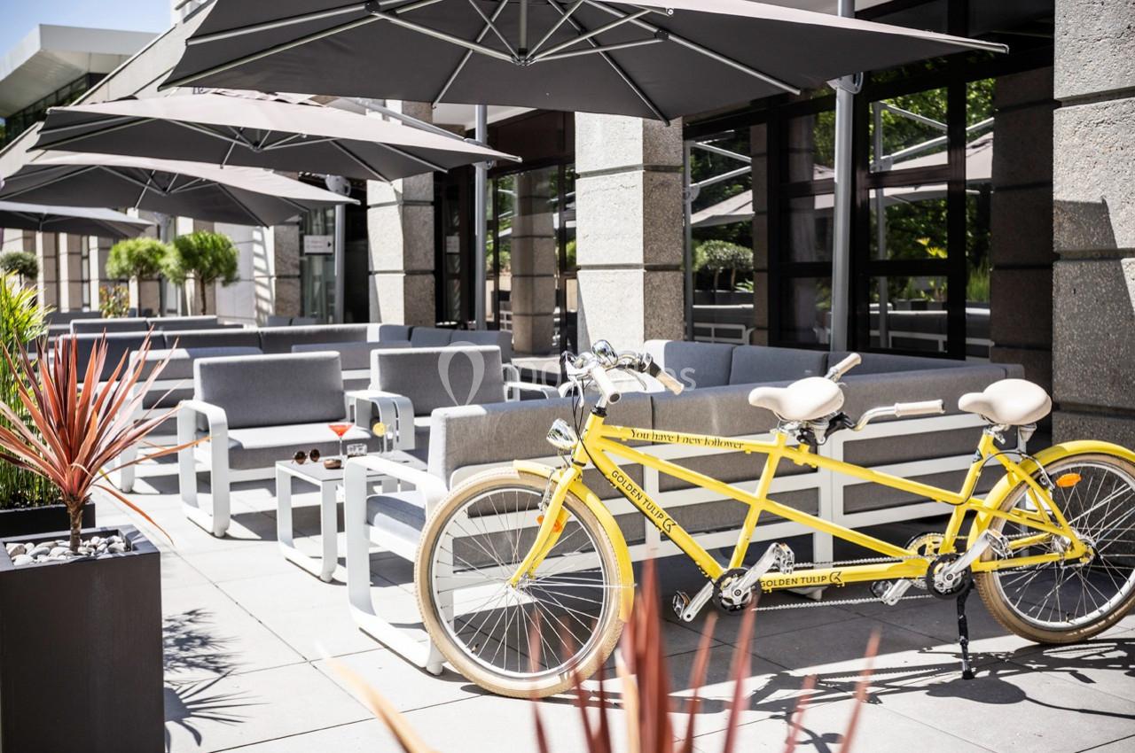 Un tandem jaune stationné sur une terrasse moderne avec des parasols et des sièges gris.