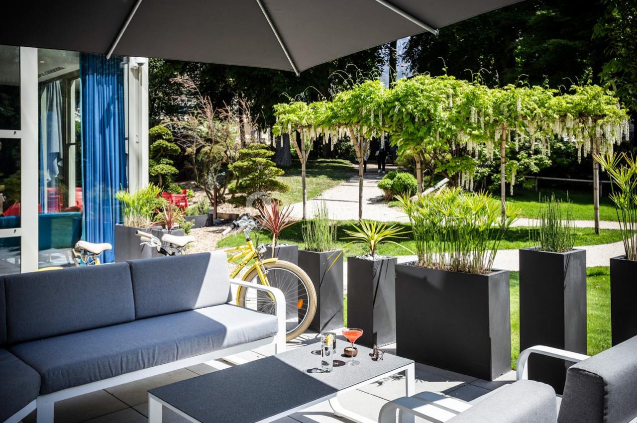 Terrasse aménagée avec canapés gris, table basse et plantes en pots, donnant sur un jardin arboré et ensoleillé.