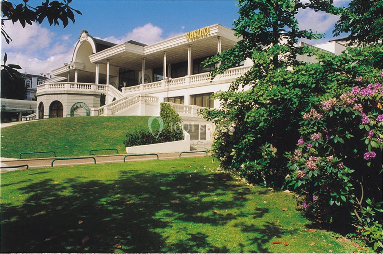 Façade d'un casino blanc entouré de verdure, avec un escalier central et des fleurs au premier plan.