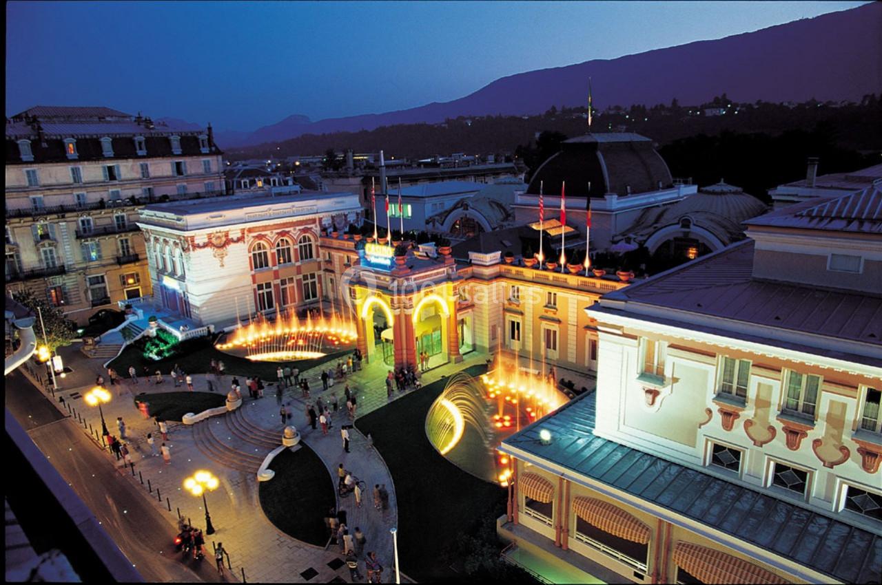 Vue nocturne d'un bâtiment historique illuminé avec des fontaines et une place animée, entouré de montagnes.