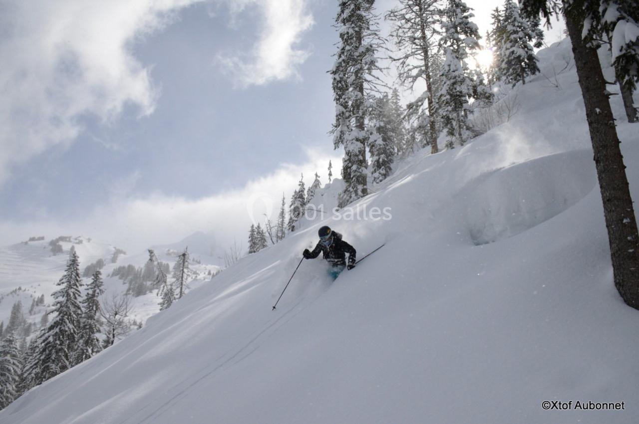 Skieur descendant une pente enneigée entourée d'arbres sous un ciel partiellement nuageux.