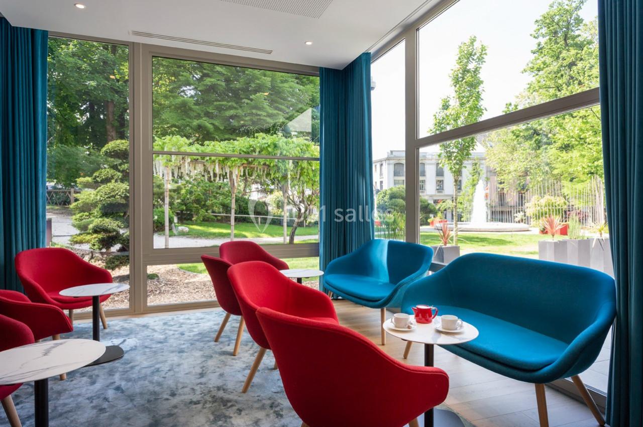 Espace salon lumineux avec fauteuils rouges et bleus, tables basses et grandes fenêtres donnant sur un jardin verdoyant.