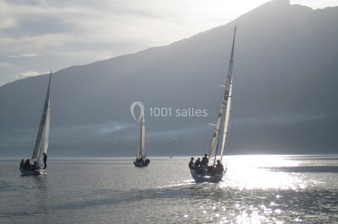 Trois voiliers naviguent sur un lac calme au lever du soleil, avec des montagnes en arrière-plan.