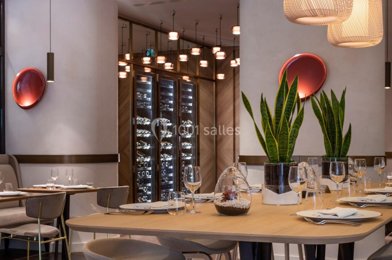 Salle de restaurant moderne avec tables dressées, plantes décoratives et cave à vin vitrée en arrière-plan.