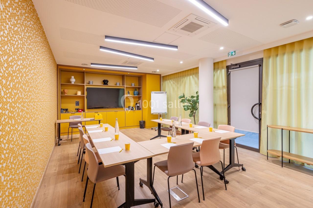 Salle de réunion lumineuse avec tables en bois, chaises colorées, tableau blanc et étagères jaunes en arrière-plan.