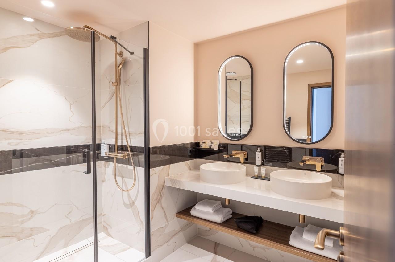 Salle de bain moderne avec douche vitrée, double vasque, miroirs ovales et finitions en marbre et laiton.