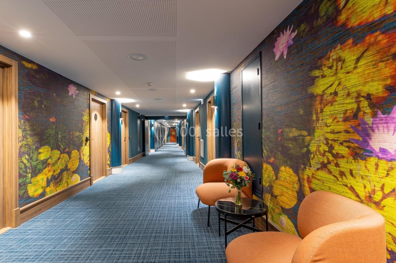Couloir d'hôtel avec moquette bleue, murs décorés de motifs floraux colorés, fauteuils orange et table avec fleurs.