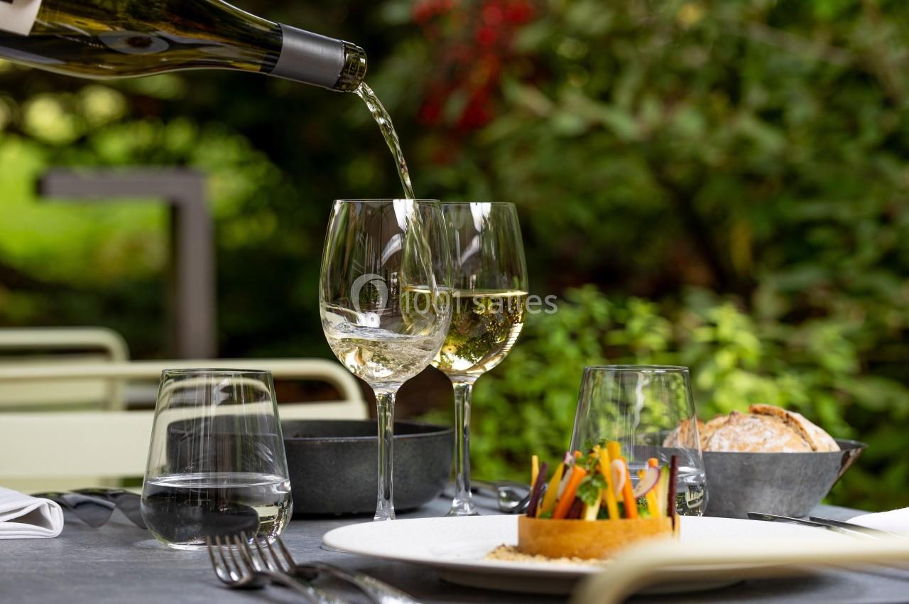 Du vin blanc est versé dans un verre sur une table dressée en extérieur avec des plats et un décor végétal en arrière-plan.