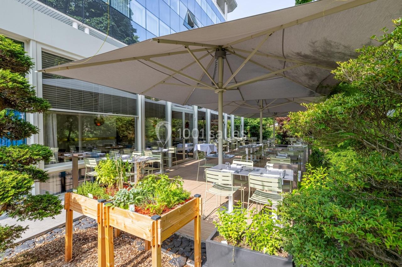 Terrasse d'un restaurant avec tables dressées, parasols et jardinières de plantes, entourée de verdure.