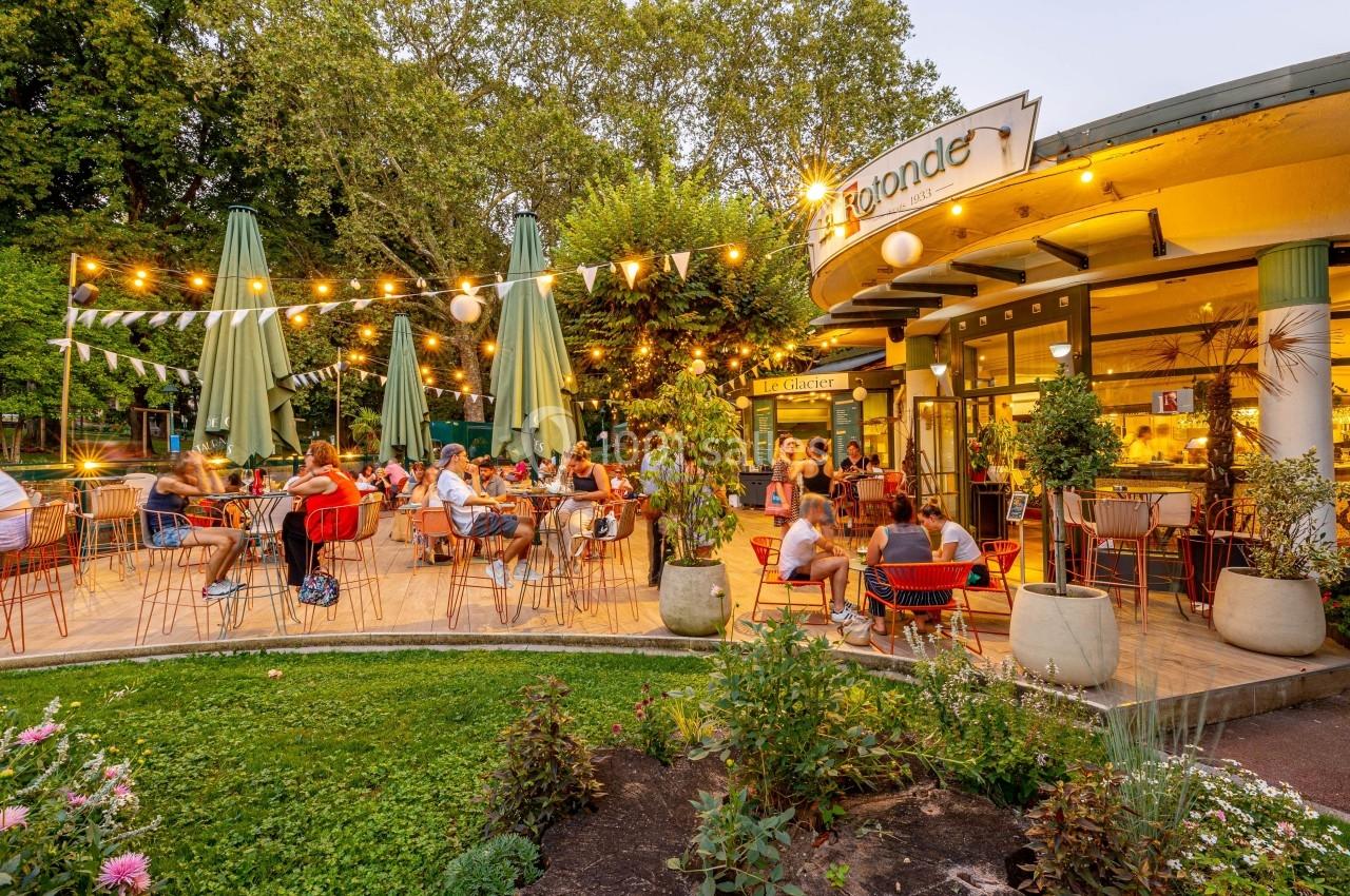 Terrasse animée d'un café-restaurant avec des tables occupées, éclairée par des guirlandes lumineuses, entourée de verdure.