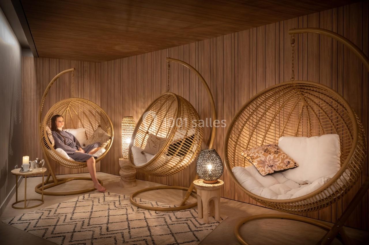 Une femme lit dans un fauteuil suspendu en rotin, dans une pièce chaleureuse aux tons boisés et éclairage tamisé.