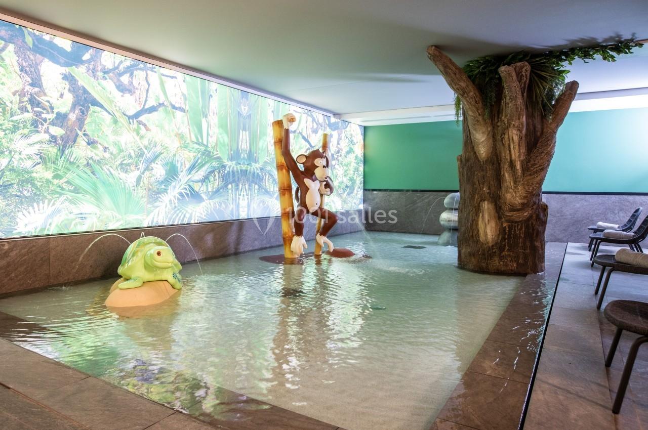 Piscine intérieure peu profonde avec décor tropical, statue de singe suspendu et tortue, entourée de chaises.