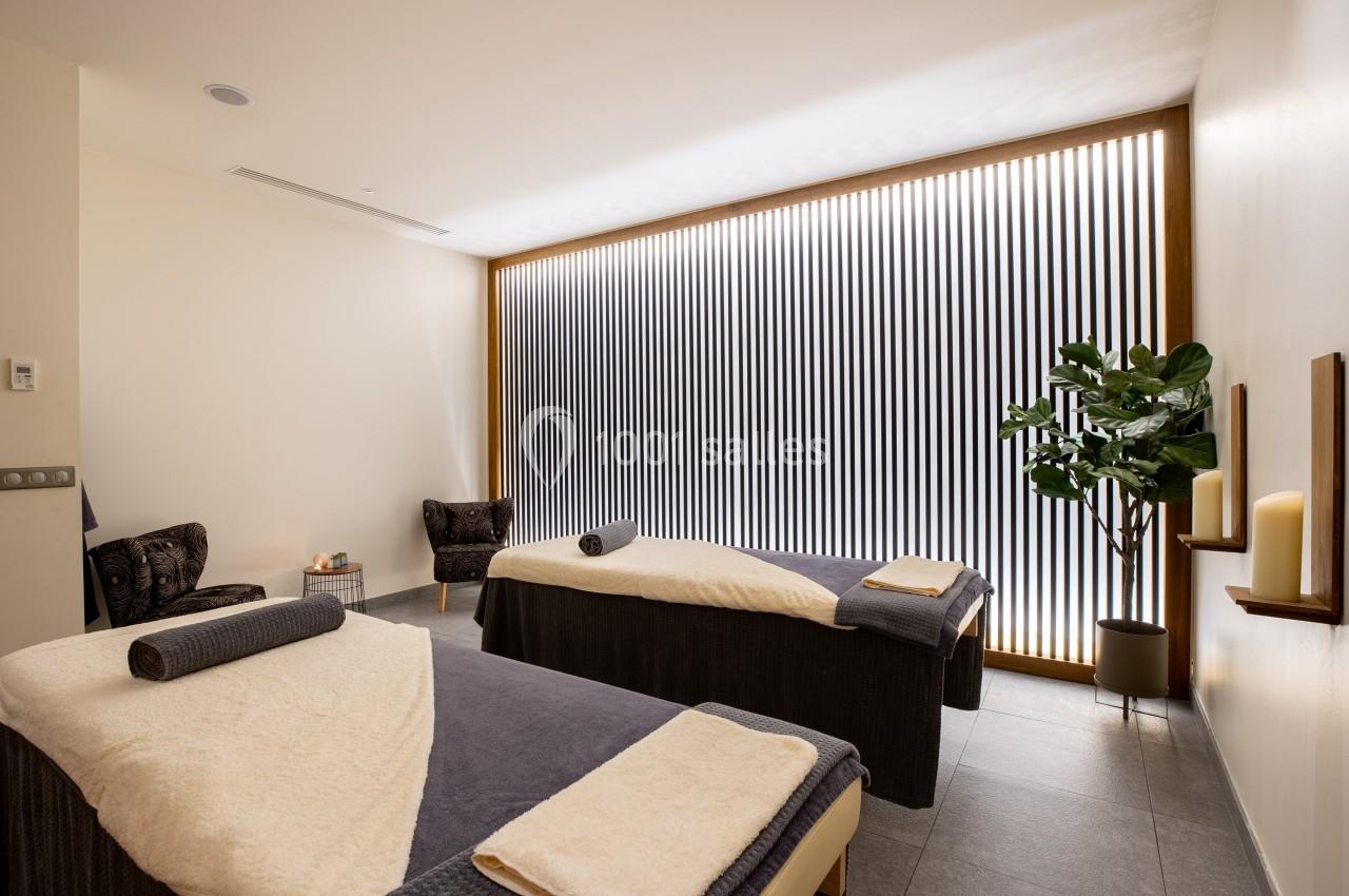 Deux tables de massage dans une salle lumineuse avec des bougies, un fauteuil et une plante en décoration.