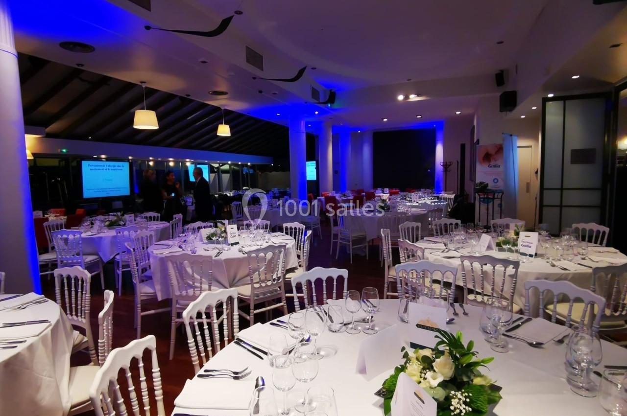 Salle de réception élégamment décorée avec des tables rondes, nappes blanches, chaises en bois et éclairage tamisé.