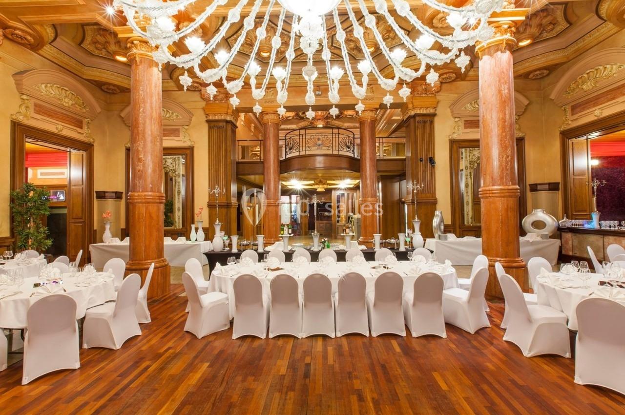 Salle de réception élégante avec tables rondes dressées, chaises blanches et grand lustre sous un plafond orné.