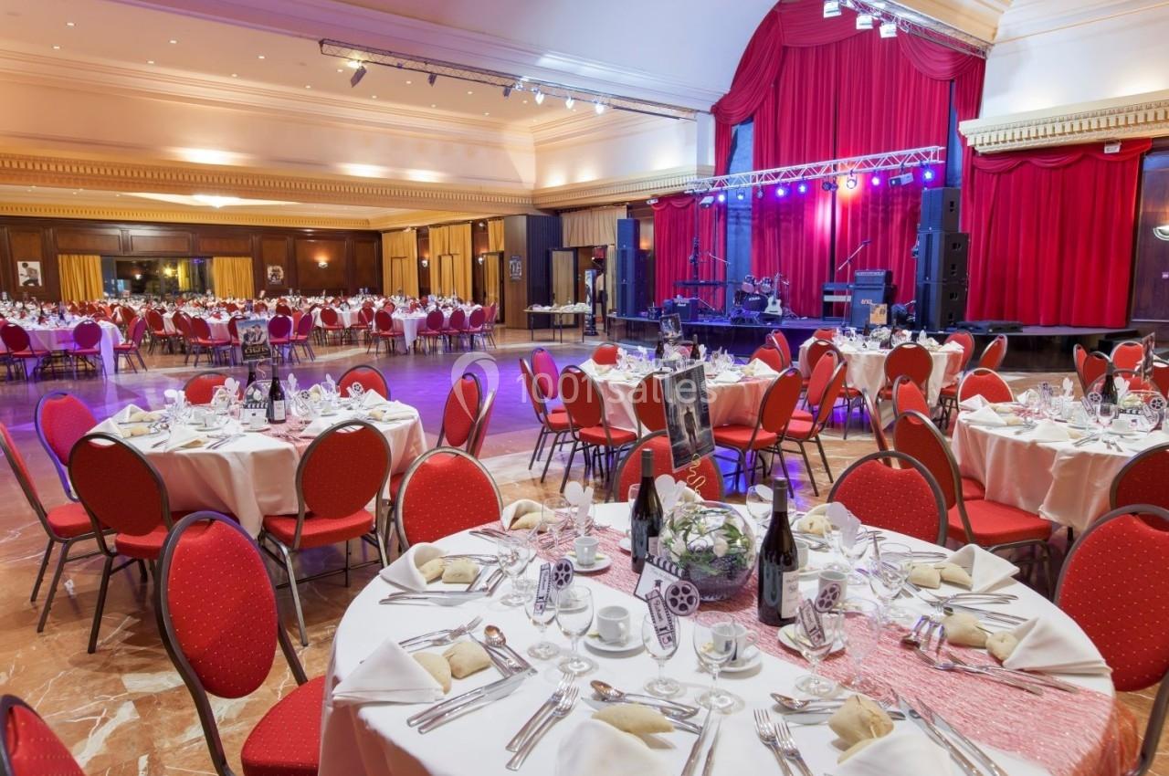 Salle de réception élégante avec tables rondes dressées, chaises rouges et scène éclairée avec rideaux rouges.