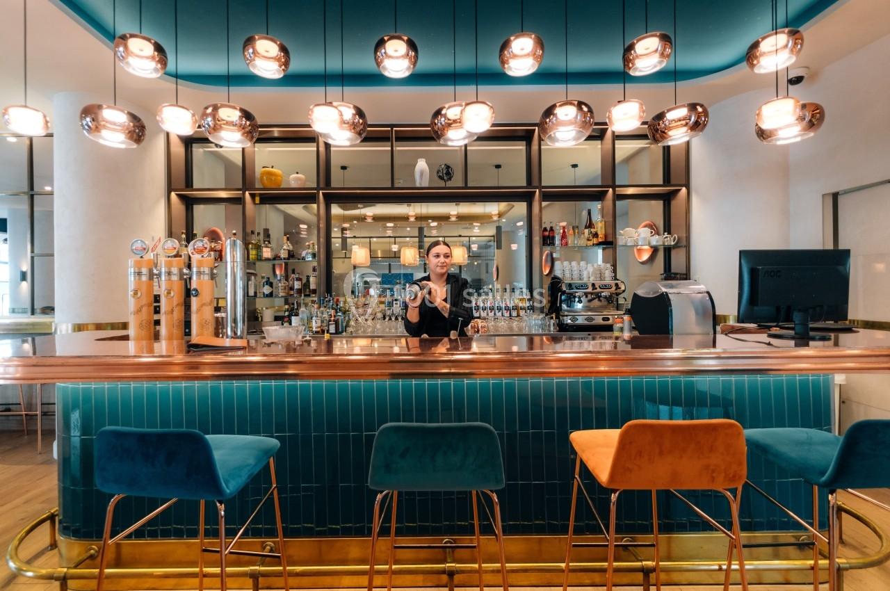 Un barman derrière un comptoir en bois dans un bar moderne avec des tabourets colorés et des luminaires suspendus.